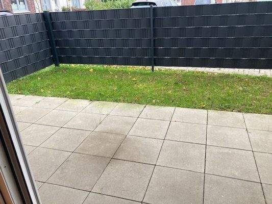 Blick von Balkontür in den Garten mit grauem Gartenzaun, helle Terrassenfliesen und Rasen