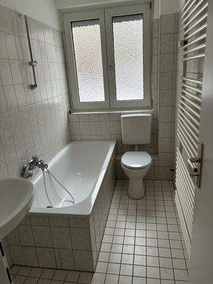 Tageslicht Badezimmer mit Badewanne und Duschvorrichtung. Weißes Stand-WC, Waschbecken und Heizkörper. Die Wände sind im Nassbereich raumhoch weiß gefliest. Der Boden ist weiß gefliest.