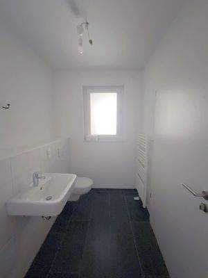 Tageslichtbad mit weißen Sanitärobjekten, weißen Wandfliesen und dunkelgrauen Bodenfliesen