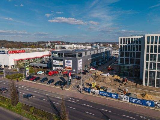 Luftaufnahme eines Gewerbegebiets mit großflächigem Einzelhandelsgebäude von Bauhaus, angrenzendem Geschäftszentrum mit Parkplätzen sowie einem modernen Bürogebäude. Im Vordergrund befindet sich eine Baustelle mit Erdarbeiten und Baggern, daneben eine bef