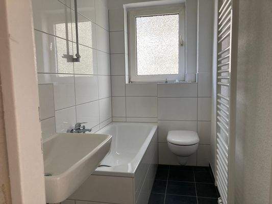 Badezimmer mit dunkelgrauen Bodenfliesen, weißen Wandfliesen, einer Badewanne und einem Fenster.