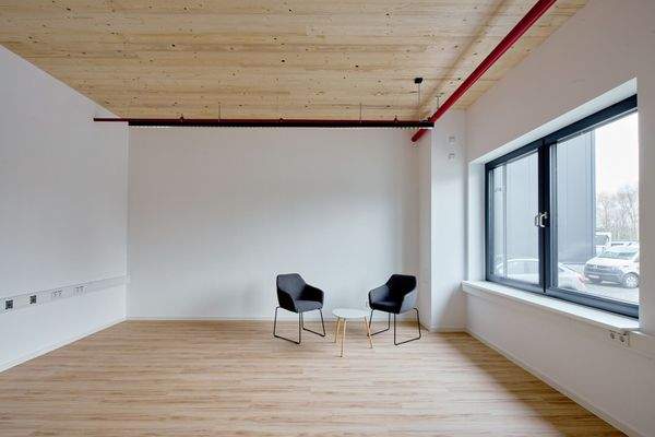 Kleiner Besprechungsbereich mit zwei schwarzen Sesseln und rundem Beistelltisch, Holzdecke mit sichtbarer Haustechnik und großem Fenster zur Straßenseite.