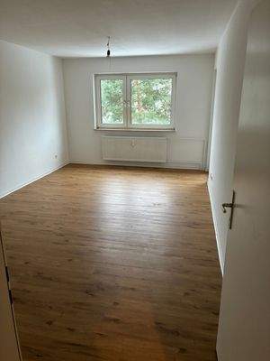 Wohnzimmer mit Fenster und Zugang zum Balkon. Wände und Decke weiß gestrichen. Brauner Vinylboden.