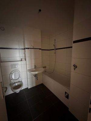 Badezimmer mit weißen Wand- und dunklen Bodenfliesen, weißem Waschbecken, WC und Badewanne. 