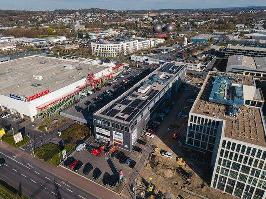 Luftaufnahme eines Gewerbegebiets mit großem Bauhaus-Markt, einem benachbarten Geschäftshaus mit Solaranlage auf dem Dach sowie einem modernen Bürogebäude mit Flachdach. Im Vordergrund eine im Bau befindliche Fläche mit Baumaschinen. Im Hintergrund sind W