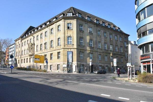 Historisches, mehrstöckiges Bürogebäude mit Sandsteinfassade an einer Straßenecke, im Vordergrund Straße mit Verkehrsschildern.