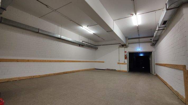 Technik- oder Lagerraum mit gefliestem Boden, weiß verputzten Wänden und sichtbarer Deckeninstallation, Tür mit Notausgangsbeleuchtung.