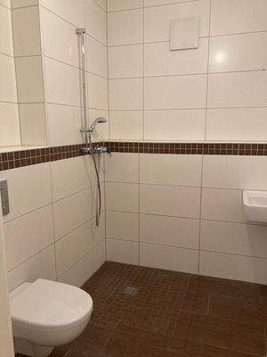  Blick in Bad mit weißen Wandfliesen, braunen Bodenfliesen Hänge-WC und ebenerdiger Dusche 