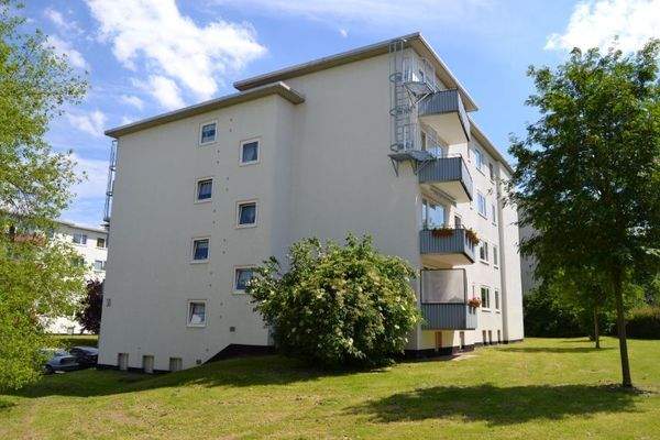 Seitenansicht eines mehrstöckigen  Wohngebäudes mit Balkonen, umgeben von Bäumen und grünen  Sträuchern 