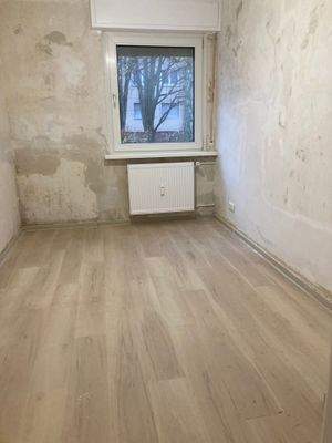 Zimmer mit Fußboden in Holzoptik, einem weißem Fenster und tapetenlosen Wänden.