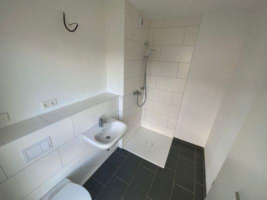 Badezimmer mit weißen Wand-WC, weißem Wandwaschbecken, barrierefreie Dusche auf grauem Fliesenboden.JPG