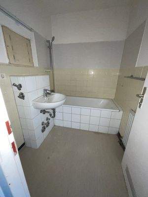 Beispielhaftes Bad mit Badewanne und Waschmaschinenanschluss, helle Wandfliesen und Linoleumboden