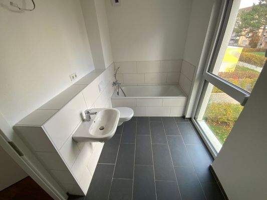 Tageslicht Badezimmer mit weißem Wand-WC, weißem Wandwaschbecken und Badewanne auf grauem  Fliesenboden.JPG