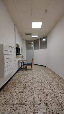 Kleiner, schmaler Raum mit Büromöbel, weißen Wänden und gefliestem Boden, an einer Wand Stromverteiler und Bildschirm.