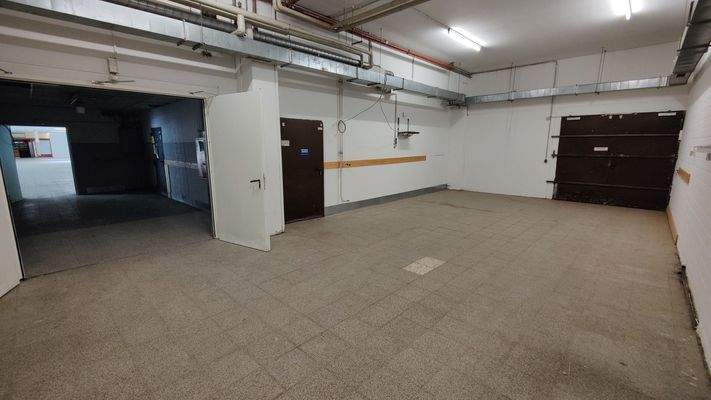 Technik- oder Lagerräumlichkeit mit offenem Durchgang, grauem Bodenbelag und sichtbarer Rohrinstallation an der Decke.