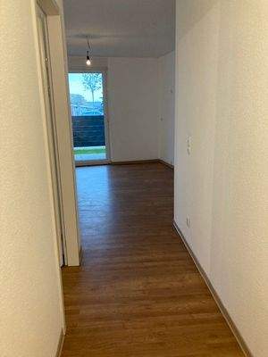 Blick Eingang in Wohnraum mit braunem PVC-Boden und weißen Wänden mit grauen ebenerdigen Fenstern 