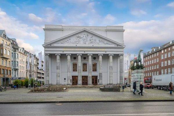 Frontansicht des klassizistischen Stadttheaters Aachen mit Säulenfassade, Giebelrelief und Inschrift. Im Vordergrund Fußgängerbereich mit Brunnenanlage und Reiterstatue.