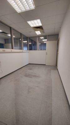 Leerer, schmaler Büroraum mit grauem Boden, weißen Wänden und Oberlichtdecken mit Leuchtstoffröhren. Rückseite mit Glastrennwänden und geöffneter Tür.