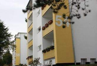 Seitenansicht eines mehrstöckigen  Wohngebäudes mit Balkonen, umgeben von Bäumen und grünen  Sträuchern 