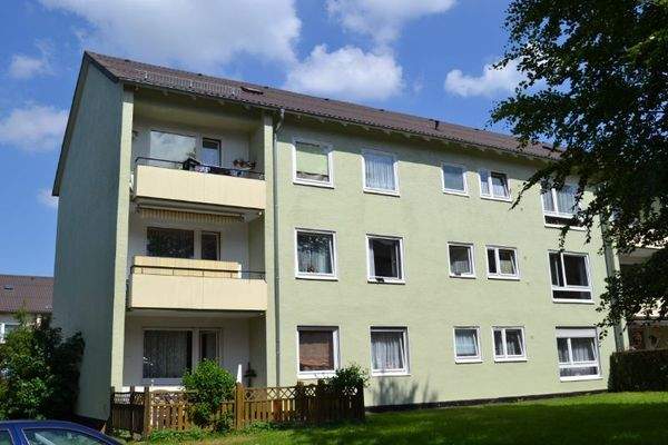 Rückansicht eines mehrstöckigen  Wohngebäudes mit Balkonen, umgeben von Bäumen und grünen  Sträuchern 