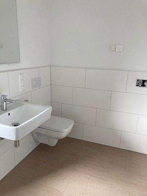 Tageslicht Bad mit Dusche mit weißem wandhängendem WC, Waschbecken und Handtuch Heizkörper. Die Wände sind im Nassbereich raumhoch und weiß gefliest. Der Boden ist hell gefliest.