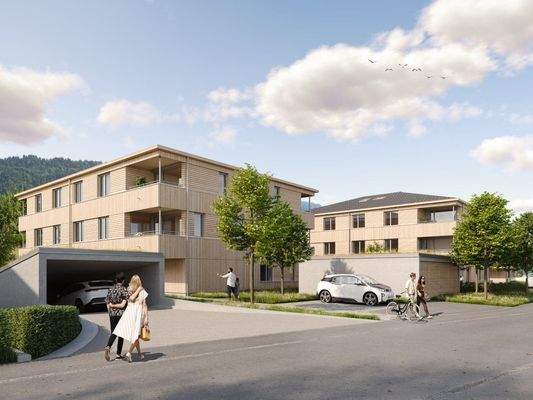 Zwei moderne Mehrfamilienhäuser mit Holzfassade und Tiefgarage, umgeben von Bäumen und Grünflächen, unter blauem Himmel in Hohenweiler (Riedstrasse).