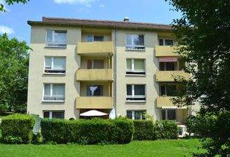 Rückansicht eines mehrstöckigen Wohngebäudes mit Balkonen, umgeben von Bäumen und grünen Sträuchern