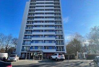 Außenansicht eines mehrstöckigen Wohngebäudes (Hochhaus), umgeben von Bäumen und Stellplätzen. 